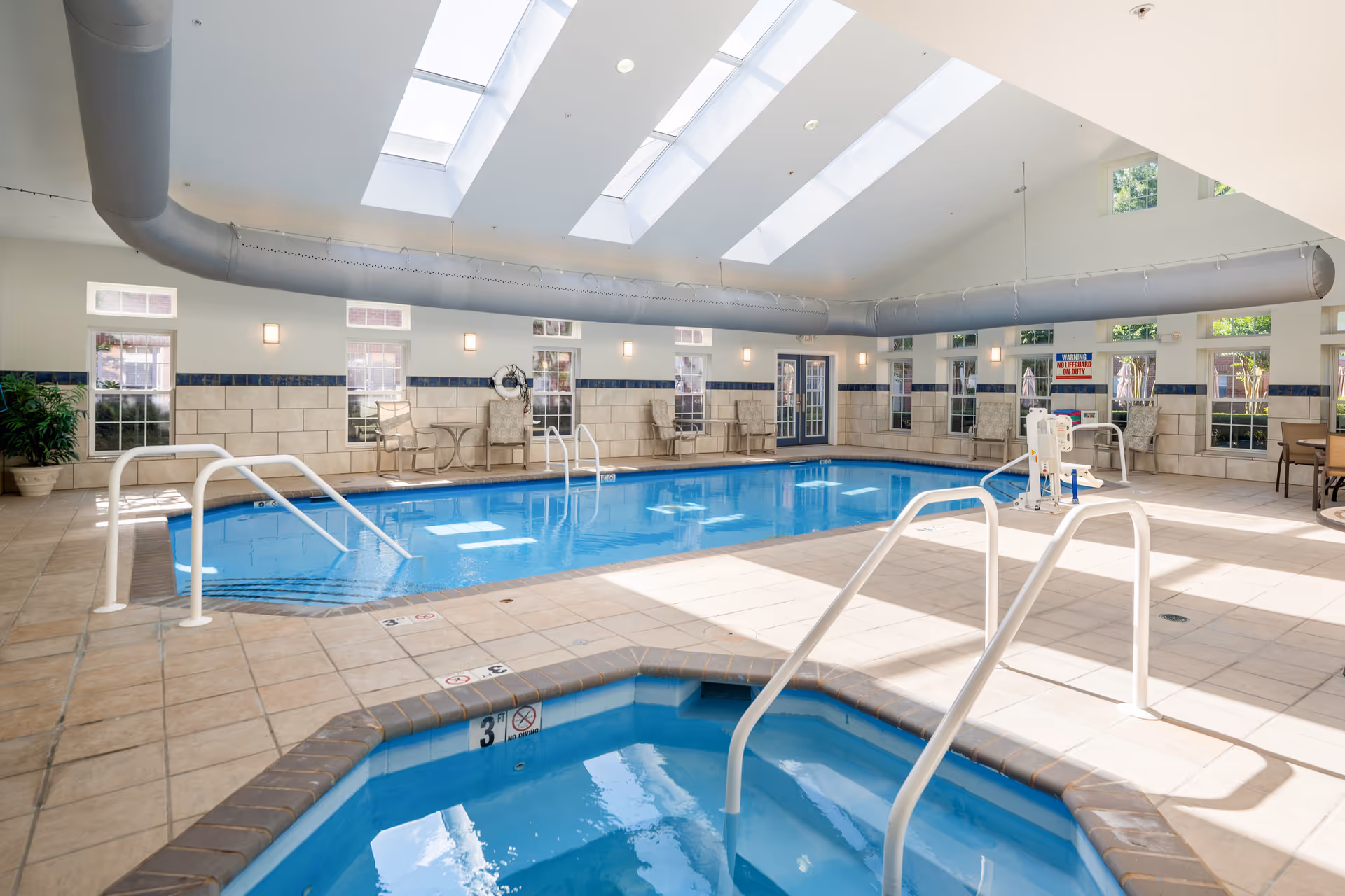 Indoor swimming pool area with a smaller hot tub in the foreground. The room has large skylights on the ceiling allowing natural light to fill the space. There are several windows along the walls, chairs, tables, and pool safety equipment visible. The pool area is clean and well-maintained with tiled flooring.