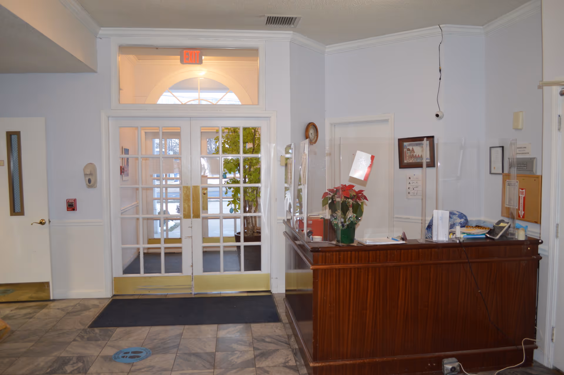 Interior view of a facility entrance area with double glass doors leading outside. There is a wooden reception desk on the right with a poinsettia plant and clear protective barriers. The floor is tiled, and there is an exit sign above the doors. A clock and some framed items are on the walls.