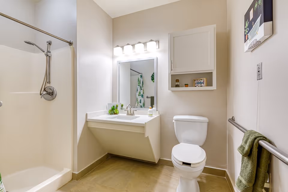 A clean and modern bathroom featuring a walk-in shower with a handheld showerhead, a white vanity with a sink and faucet, a large mirror with four lights above it, a white toilet, a wall-mounted cabinet with decorative items, a towel rack with a green towel, and a small framed picture on the wall.