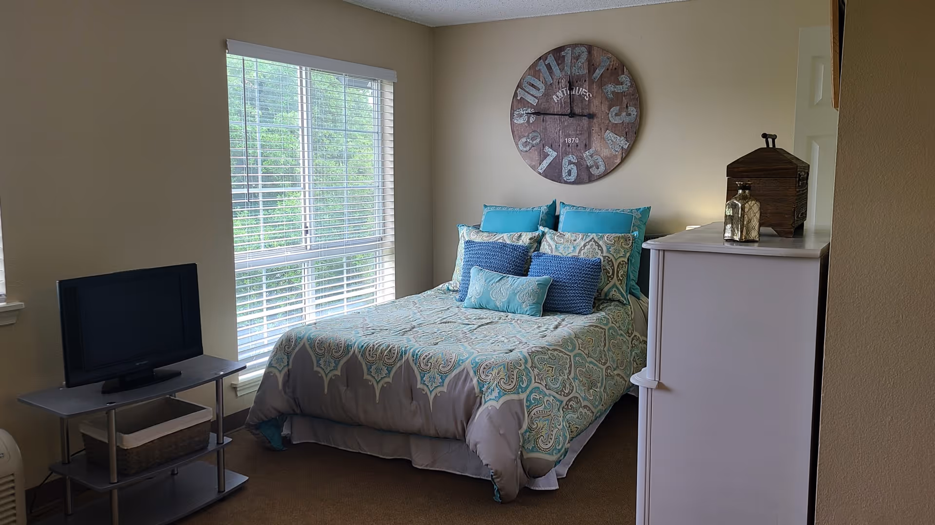 A bedroom with a bed covered in a patterned comforter and multiple pillows in shades of blue and teal. A large round wooden clock hangs on the wall above the bed. To the left of the bed is a window with white blinds, and a small TV sits on a gray stand with a basket underneath. On the right side, there is a white dresser with decorative items on top.