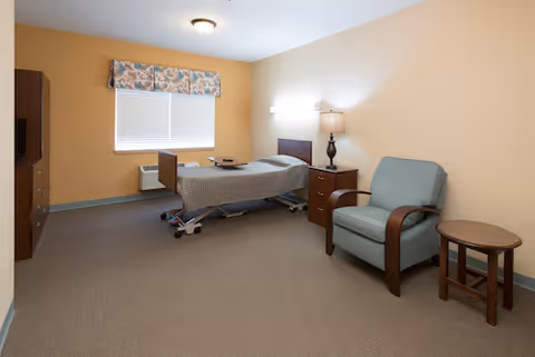 A senior care private bedroom with an adjustable hospital bed, bedside table and lamp, armchair, small table, and a window.