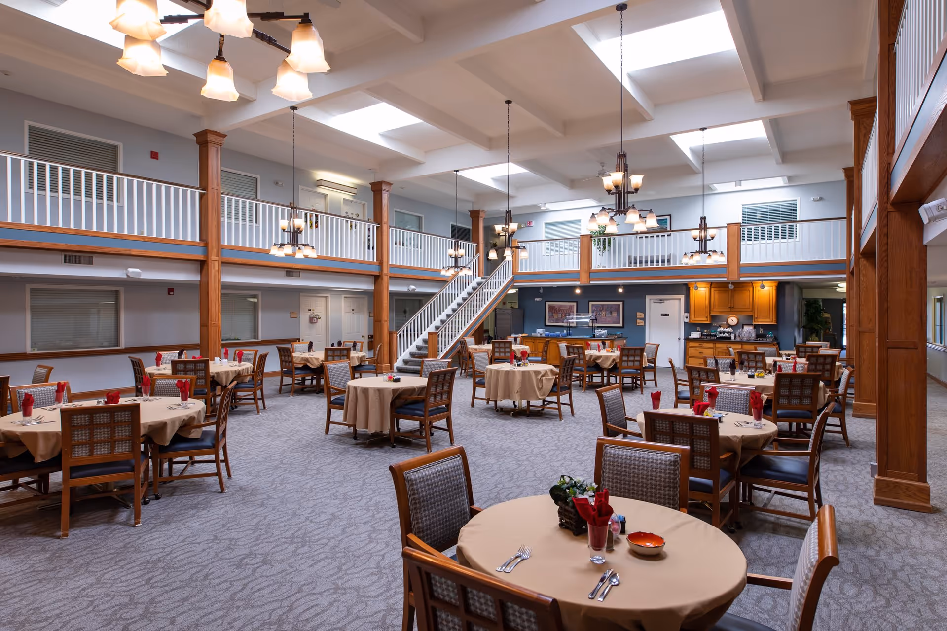 Spacious dining room with multiple round tables covered with beige tablecloths, each set with utensils, napkins, and small centerpieces. The room features a high ceiling with skylights, wooden columns, a staircase leading to an upper balcony, and warm lighting fixtures hanging from the ceiling. The walls are light-colored with large windows and a kitchen area visible in the background.