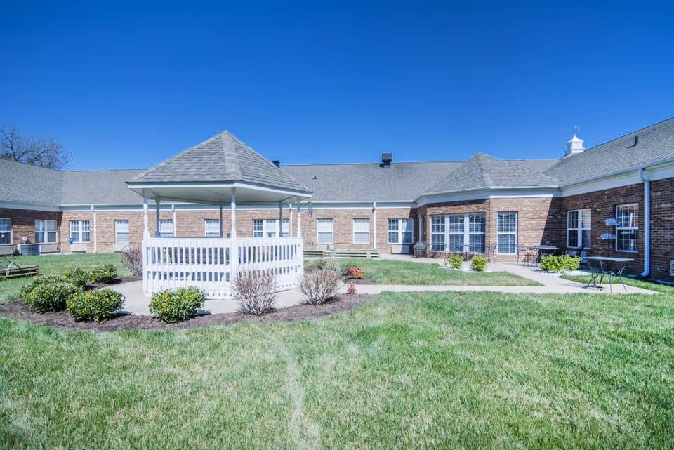 Outdoor courtyard area of a senior living facility with a white gazebo in the center surrounded by green grass, small bushes, and a brick building with multiple windows in the background under a clear blue sky.