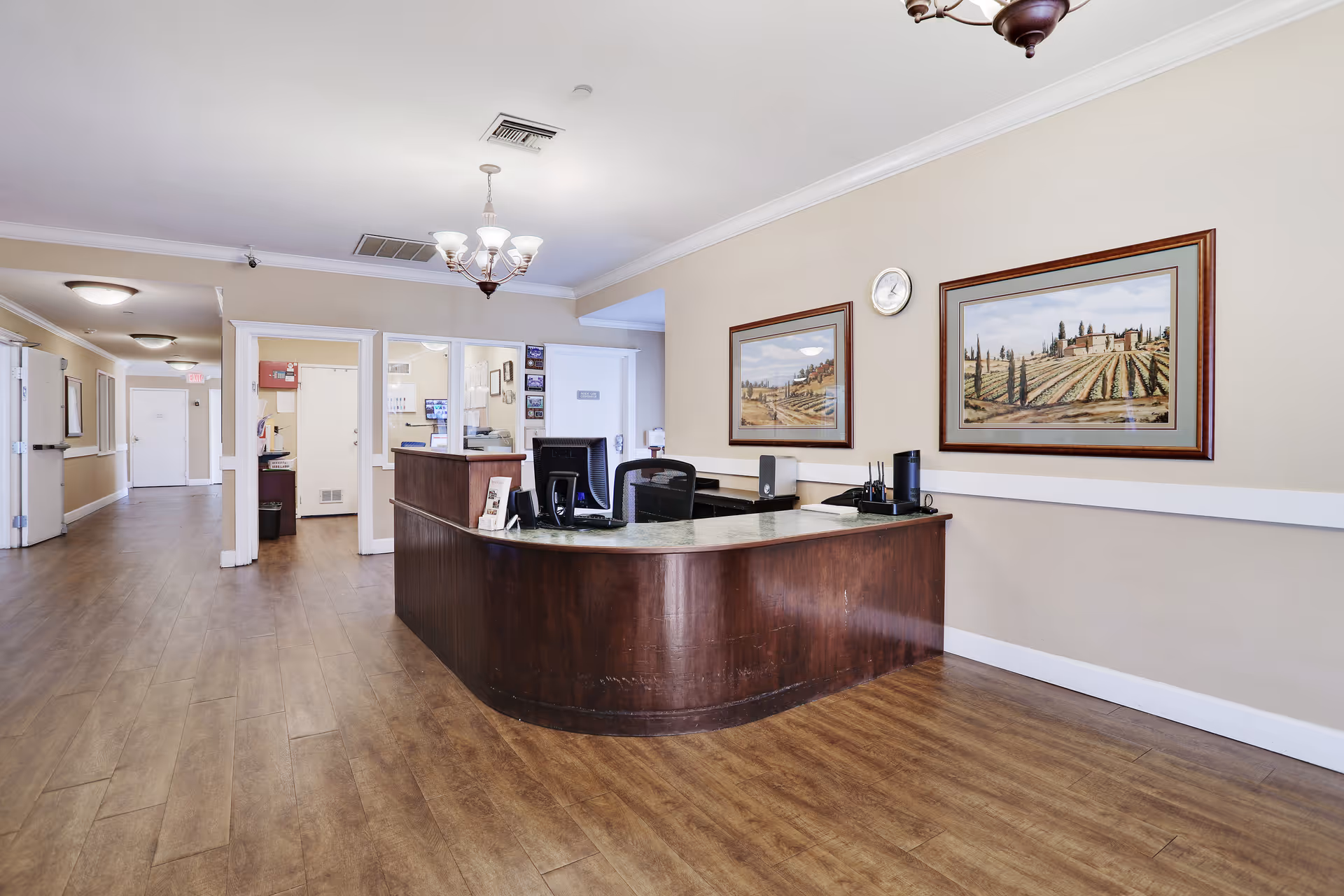 Reception desk and hallway inside an assisted living facility with framed landscape paintings on the wall.
