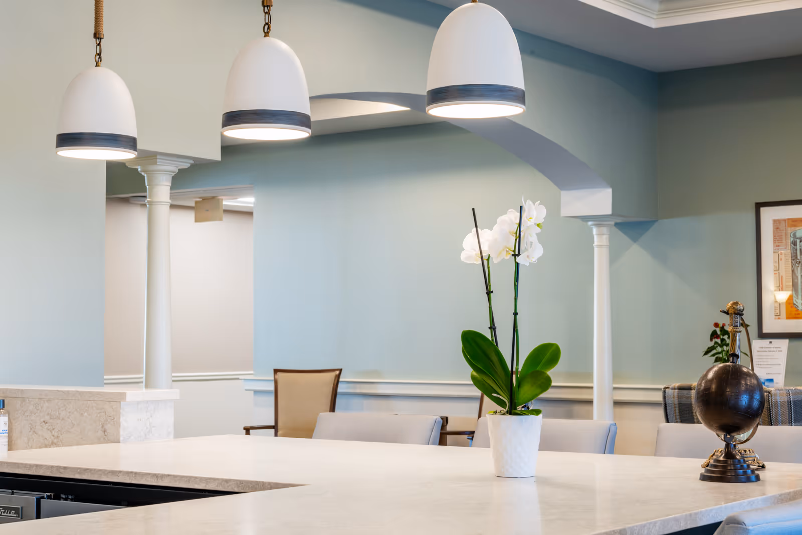 Interior view of a common area with a marble countertop, three hanging pendant lights, a white orchid plant in a pot, chairs, and a decorative globe on the counter. The walls are painted light blue with white trim and columns.