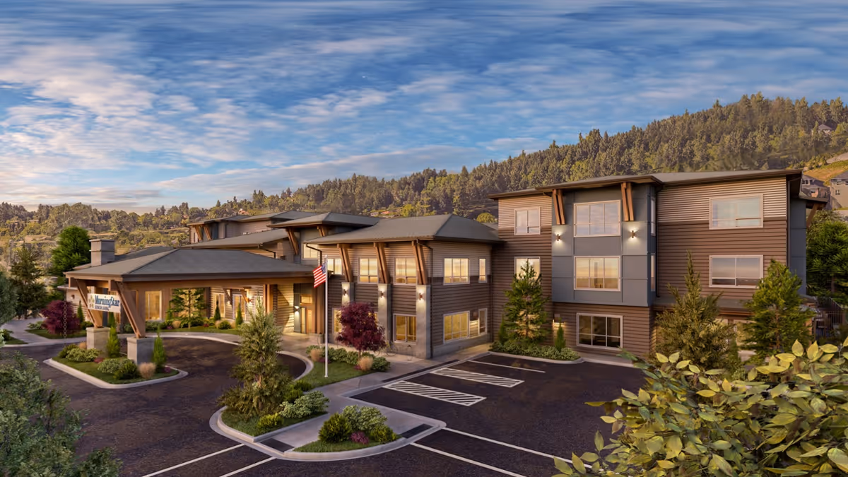 Exterior view of MorningStar Assisted Living & Memory Care of Happy Valley, a modern three-story building surrounded by landscaped greenery and trees, with a driveway and parking spaces in front, set against a backdrop of forested hills under a partly cloudy sky.