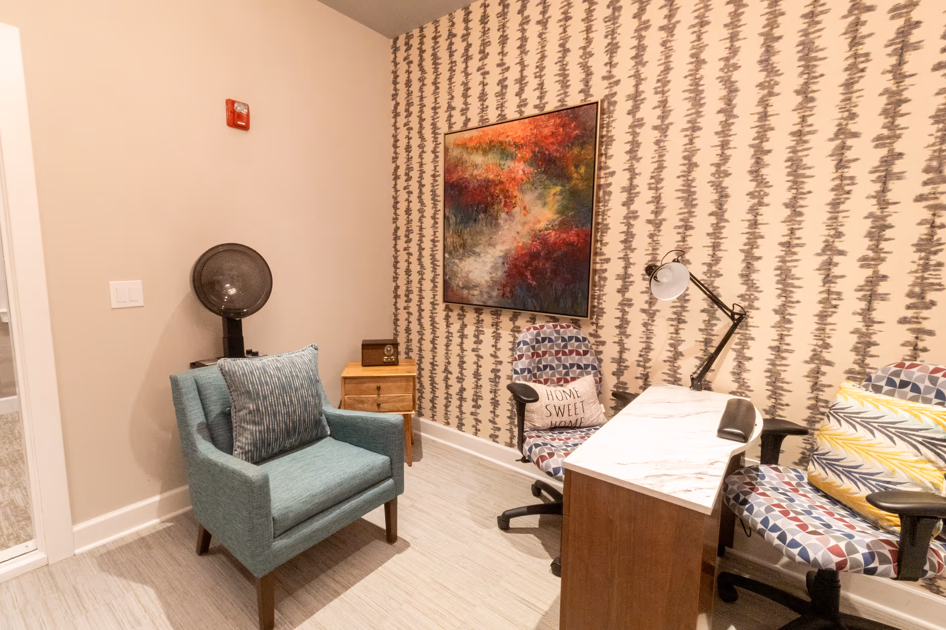 A cozy room with a patterned wallpaper wall featuring a colorful abstract painting. The room contains a teal armchair with a matching pillow, a small wooden side table with a vintage radio, and a desk with a marble-like surface. Two office chairs with geometric patterned upholstery and decorative pillows are positioned at the desk, one with a 'Home Sweet Home' pillow. A black adjustable desk lamp is attached to the desk.