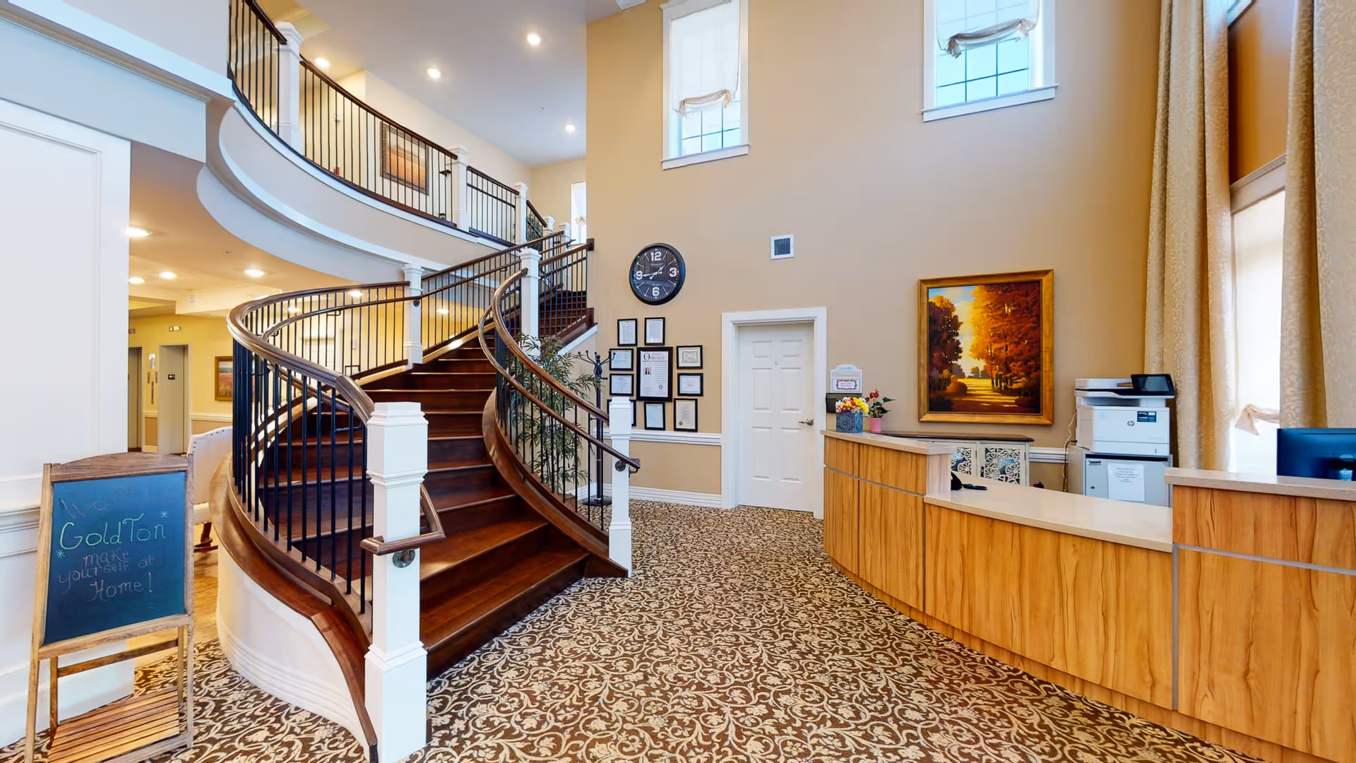 Interior view of a senior living facility lobby featuring a curved wooden staircase with black railings, a patterned carpet, a wooden reception desk with a printer, a wall clock, framed certificates, a painting of a tree-lined path, and a chalkboard sign that reads 'GoldTon make yourself at Home!'