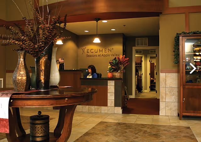 Reception area of Ecumen Seasons at Apple Valley with a woman seated behind the front desk. The area features a round wooden table with decorative vases and plants, tiled flooring, warm lighting, and a display cabinet on the right side.