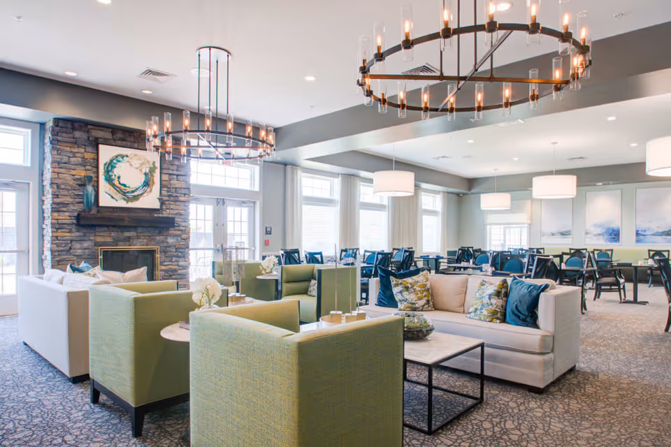 Bright, open senior living common room with sofas and green armchairs arranged around a coffee table, a stone fireplace, and dining tables in the background.