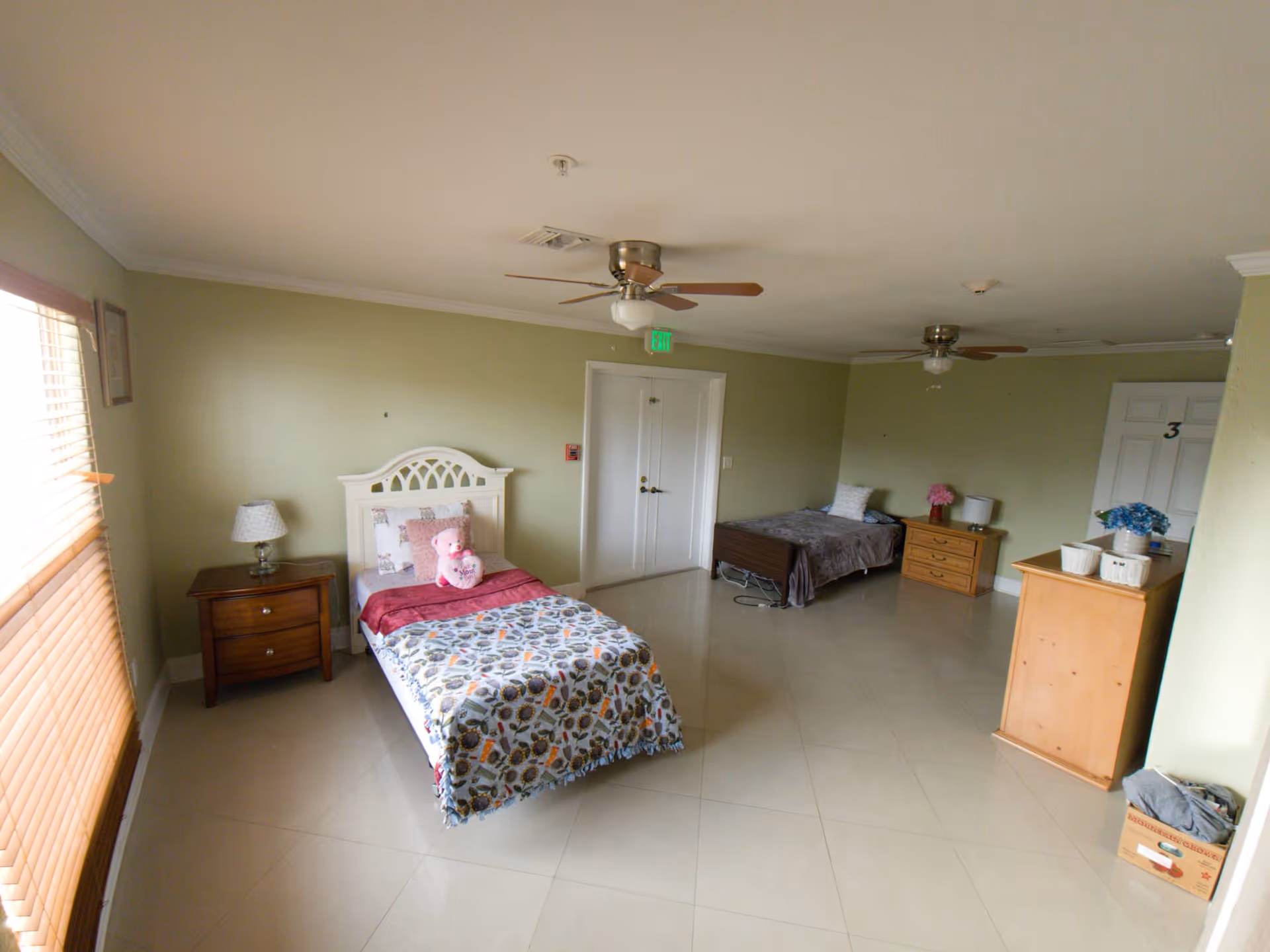 A spacious bedroom in an assisted living facility with two single beds, each with bedside tables and lamps. One bed has a colorful blanket and a pink teddy bear, while the other has a gray blanket and a pink flower vase on the nightstand. The room has light green walls, tiled floors, ceiling fans, and a window with wooden blinds letting in natural light.
