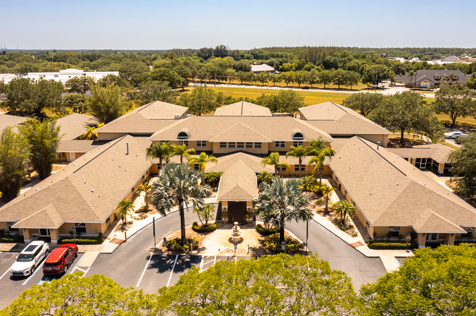 Aerial view of a single-story senior living facility with a beige roof and a U-shaped layout surrounded by trees and greenery. The entrance features a covered driveway with palm trees and a small fountain in the center. Several cars are parked in the parking lot adjacent to the building.