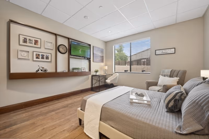 Well-lit furnished bedroom with a made bed, armchair, desk, wall-mounted TV and decorative shelving by a window.