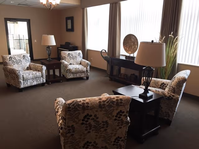A cozy living room area with four floral patterned armchairs arranged around two dark wooden side tables with lamps. There is a console table against the window with decorative items and tall plants beside it. The room has large windows with vertical blinds and beige walls.