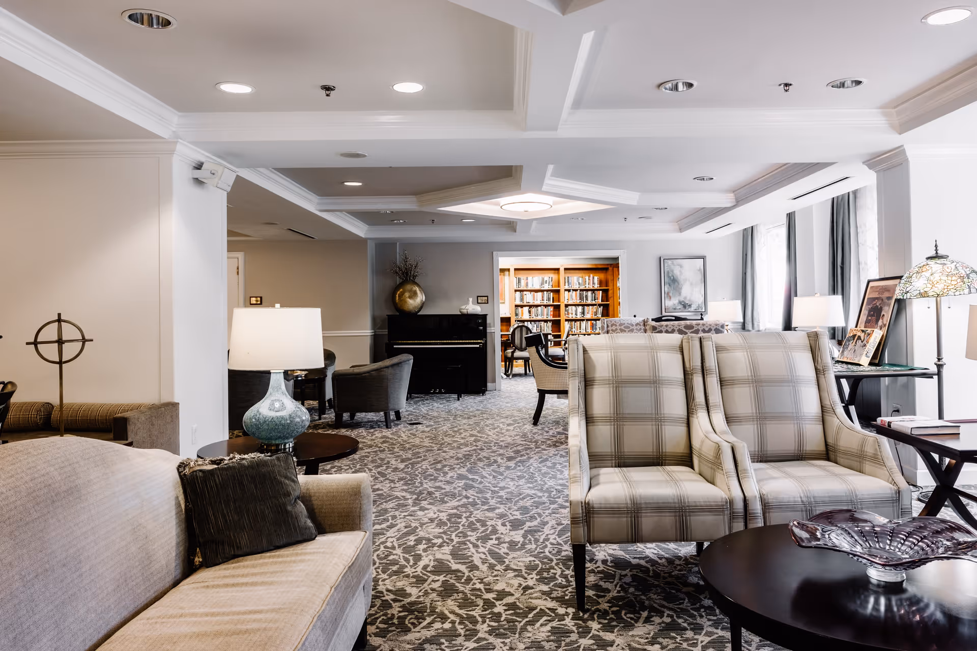 A spacious and well-lit common area in Thomas House Retirement Community featuring comfortable seating including a beige sofa and two plaid armchairs. The room has a patterned carpet, several lamps, and a piano against the far wall. In the background, there is a doorway leading to a library with bookshelves and chairs.