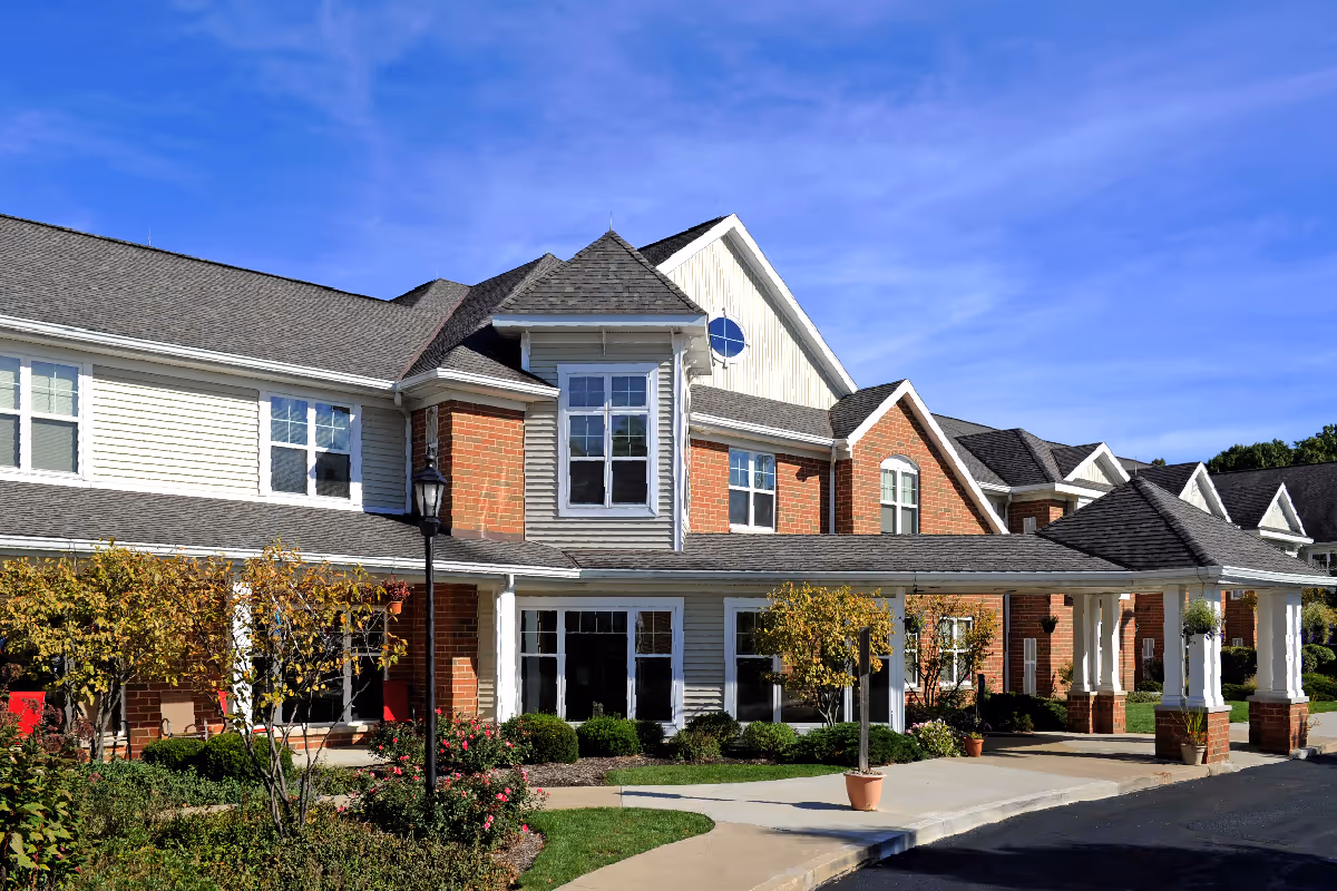 Front exterior of a two-story brick and siding senior living building with a covered entrance, landscaping, and a clear blue sky.