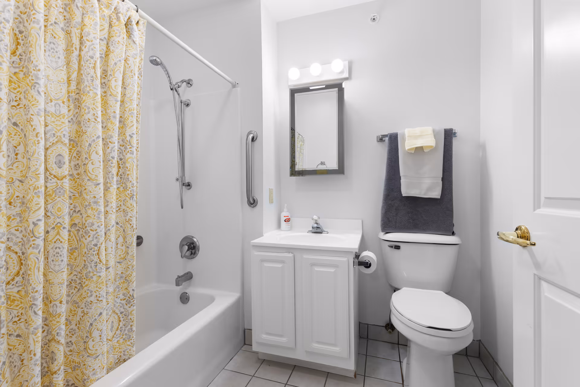Small clean bathroom with a bathtub and yellow patterned shower curtain, white vanity with mirror, and a toilet with towels.