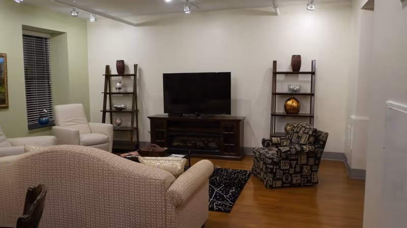 A cozy living room with a beige patterned sofa, a white armchair, and a patterned armchair arranged around a black area rug with white designs. A flat-screen TV sits on a dark wooden TV stand against a light-colored wall, flanked by two wooden ladder-style shelves holding decorative vases and bowls. The room has wooden flooring and a window with blinds on the left side.