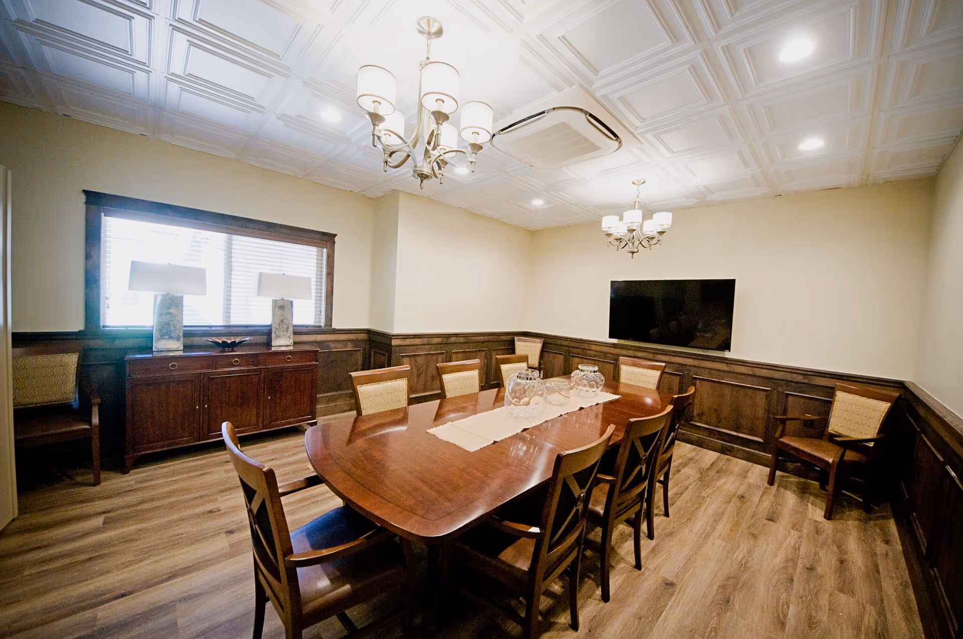 A well-lit dining room with a long wooden table surrounded by eight chairs. The room features wood paneling on the lower walls, a wooden sideboard with two lamps, a large window with blinds, two chandeliers hanging from a decorative ceiling, and a flat-screen TV mounted on the wall.