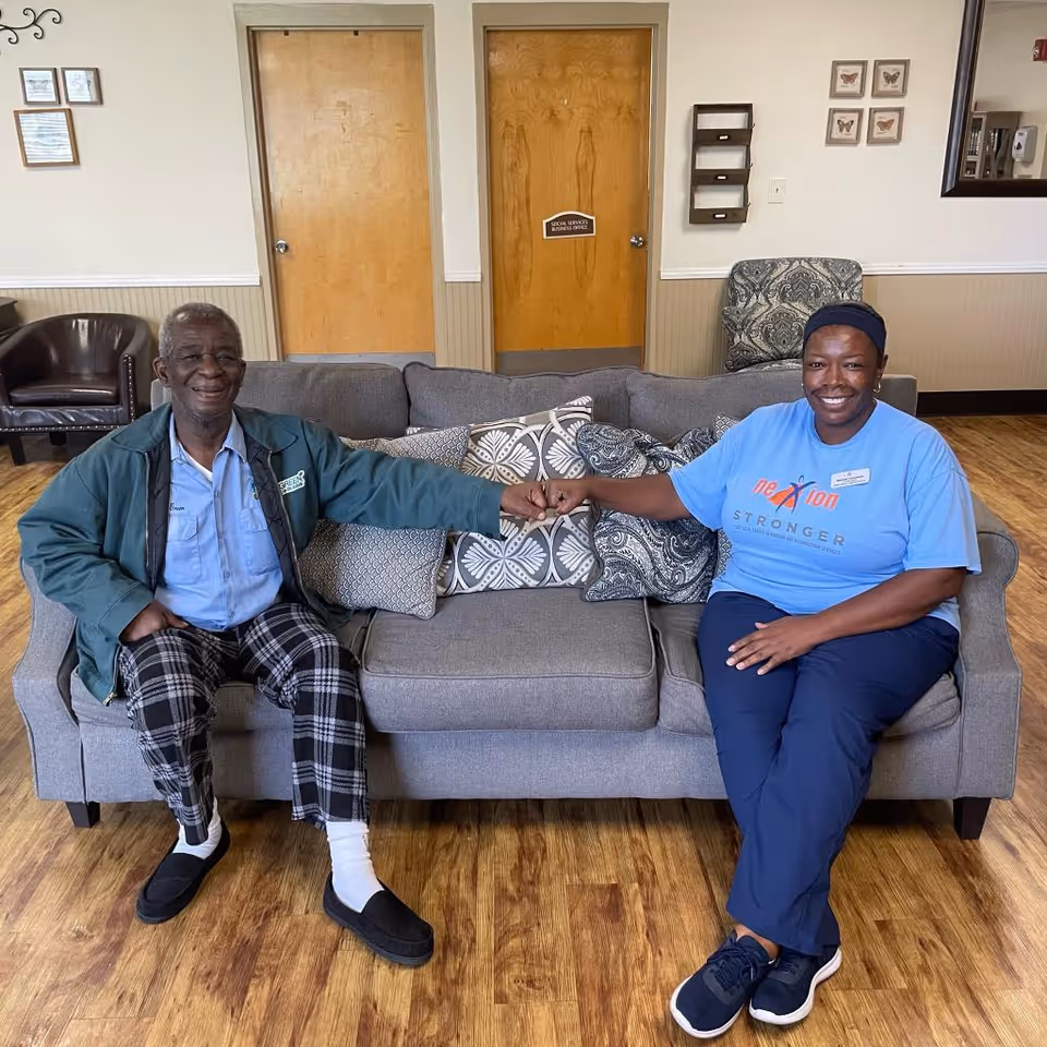 An elderly man and a woman sitting on a gray couch in a room with wooden floors. They are smiling and fist bumping each other. Behind them are two wooden doors, one labeled 'Social Services Nursing Office', a brown leather chair, a patterned armchair, and some framed butterfly pictures on the wall.