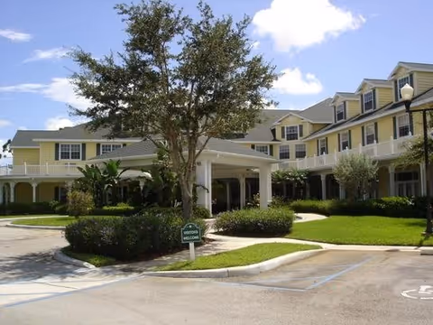 Exterior view of a large two-story senior living facility building with yellow siding and white trim. The building has multiple windows and dormers, a covered entrance with a driveway, landscaped greenery including bushes and a tree, and a clear blue sky with some clouds.