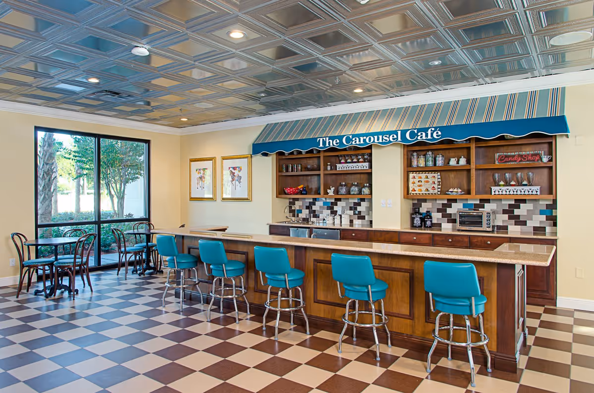 Interior view of The Carousel Café in Volante Senior Living of St. Petersburg featuring a checkered floor, a counter with five turquoise bar stools, wooden shelves with jars and decorations, a toaster oven, and two small tables with chairs near a large window showing outdoor greenery.