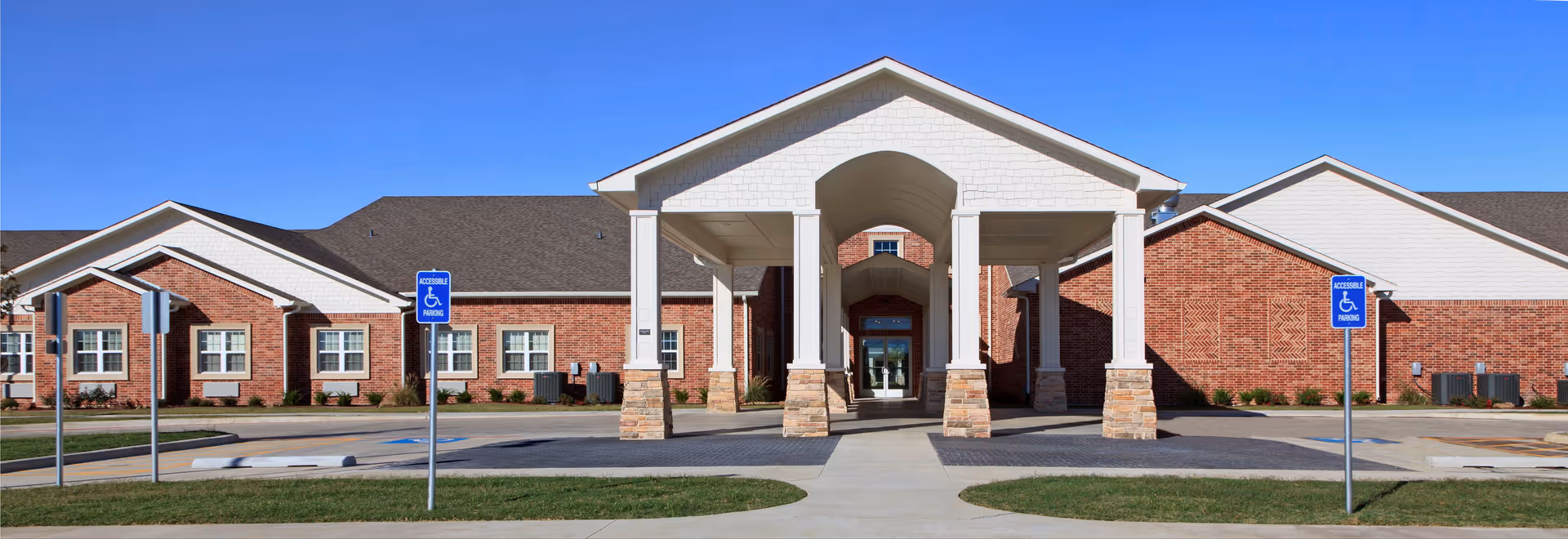 Front exterior view of Autumn Wind Assisted Living facility featuring a large covered entrance with white columns and stone bases, red brick walls, multiple windows, and accessible parking spaces with blue signs in front.