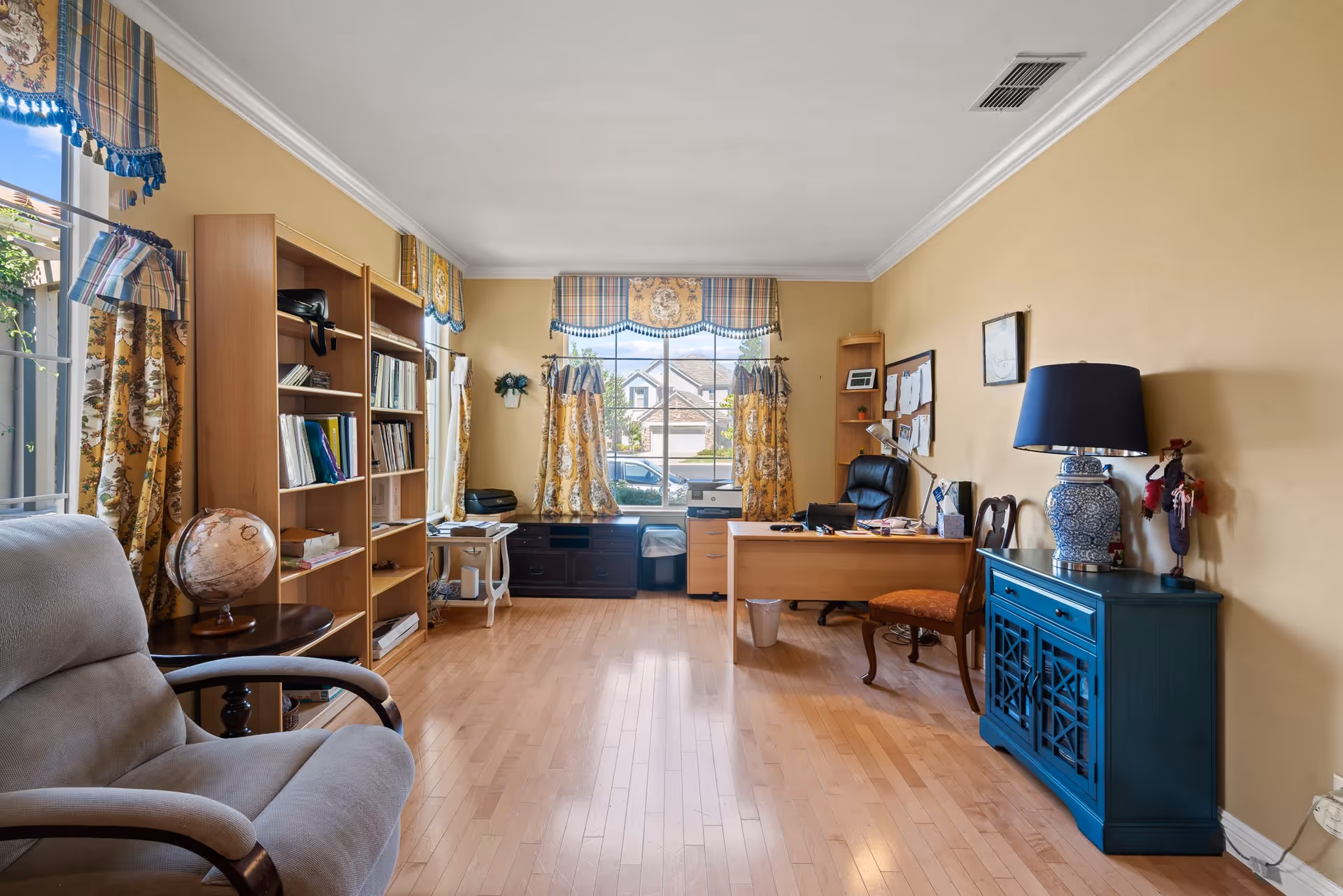 A well-lit office room with light wooden flooring and beige walls. The room features a large window with patterned yellow and blue curtains, a wooden desk with office supplies and a black office chair, a blue cabinet with a decorative lamp, a wooden bookshelf filled with books, and a comfortable armchair. The room has a cozy and organized atmosphere.