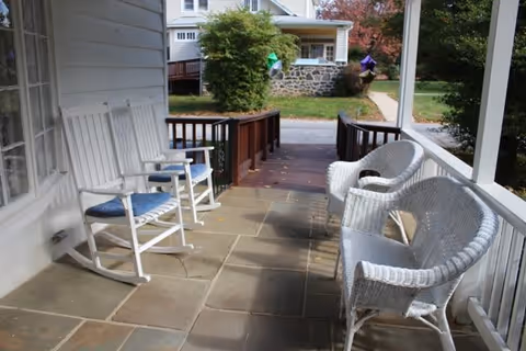 A covered porch with white rocking chairs and wicker chairs arranged along the sides. The porch overlooks a street with houses and trees in the background.