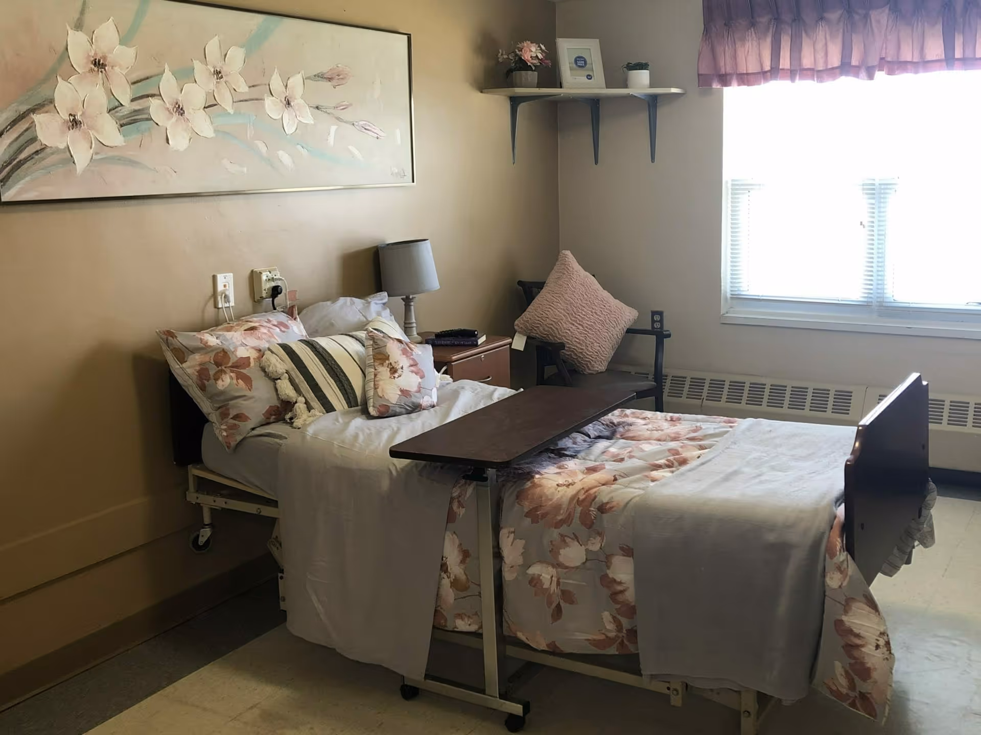 A furnished senior living bedroom with an adjustable bed dressed in floral bedding, an overbed table, bedside lamp, chair and a window.