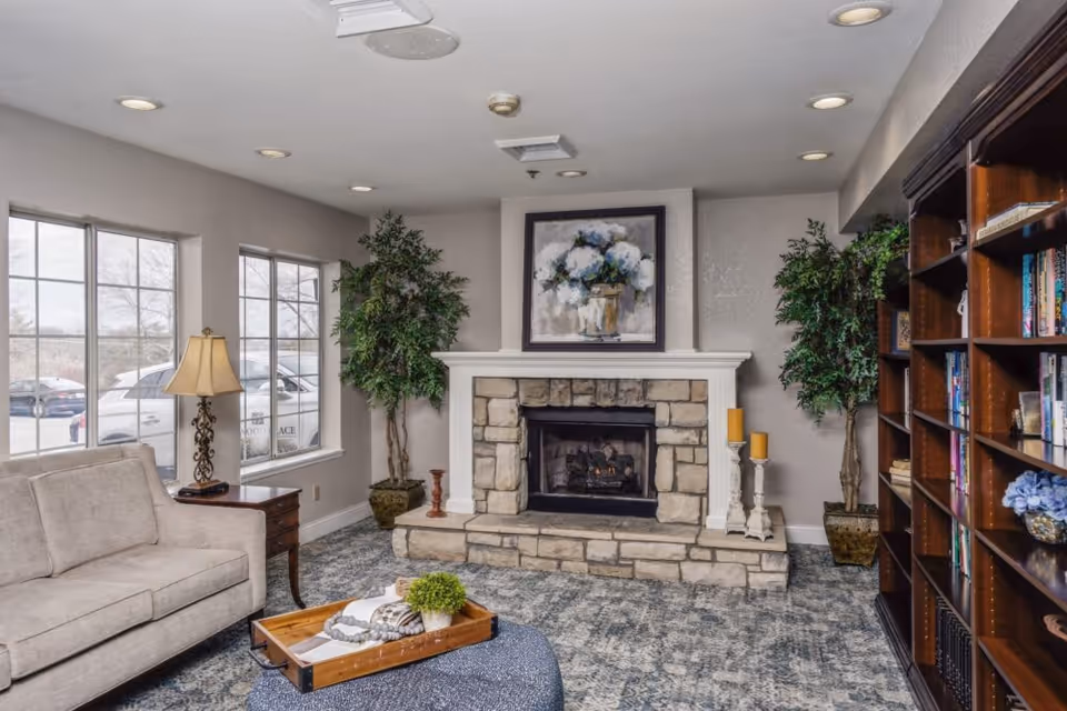 A cozy living room with a stone fireplace centered on the far wall, flanked by two potted plants. Above the fireplace hangs a painting of white flowers in a vase. To the left, there is a beige sofa next to a wooden side table with a lamp. On the right side, a large wooden bookshelf filled with books and decorative items. The room has large windows letting in natural light and a patterned carpet covering the floor.
