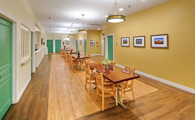 A long hallway with wooden flooring and yellow walls featuring several tables with chairs arranged along the right side. The hallway has green doors and framed pictures on the walls, with ceiling lights hanging above the tables.