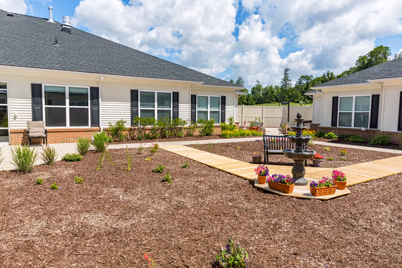 Outdoor courtyard area at StoryPoint Rockford featuring a central water fountain surrounded by flower pots and a bench. The courtyard is bordered by single-story buildings with white siding, black shutters, and large windows. Mulched garden beds with small plants and shrubs are visible, along with a wooden walkway leading to the fountain. The sky is partly cloudy with blue patches.