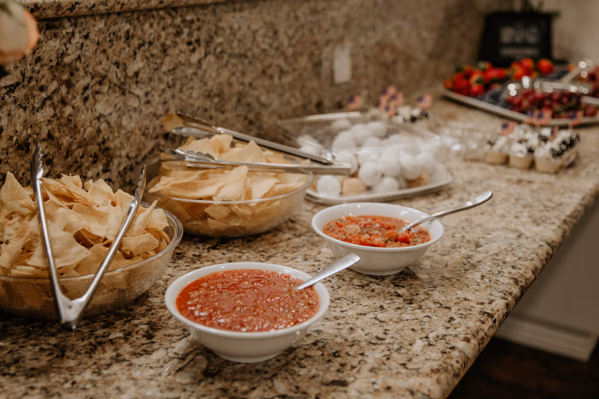 Bowls of tortilla chips and two bowls of salsa on a granite countertop with trays of fruit and desserts in the background.