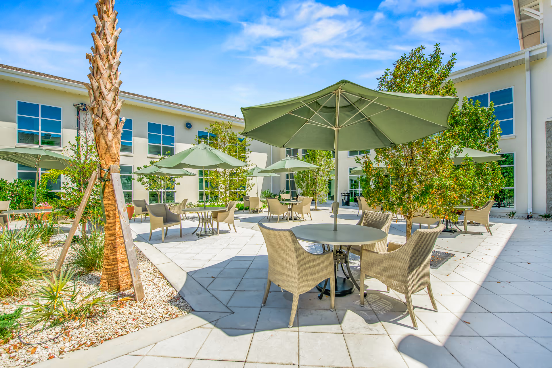 Outdoor patio area at Pines of Sarasota Health Care, LLC with round tables, wicker chairs, and large green umbrellas. The space is surrounded by a two-story building with large windows, palm trees, and other greenery under a blue sky with some clouds.