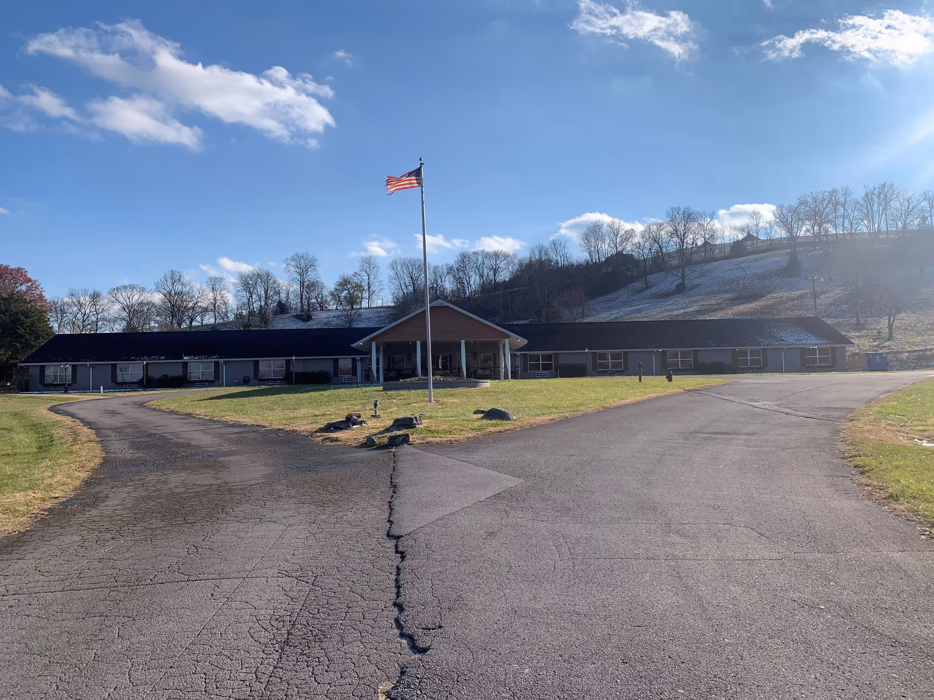 Exterior view of a single-story assisted living facility building with a peaked entrance and an American flag on a flagpole in front. The building is surrounded by a paved driveway and grassy areas, with leafless trees and a hill in the background under a partly cloudy blue sky.