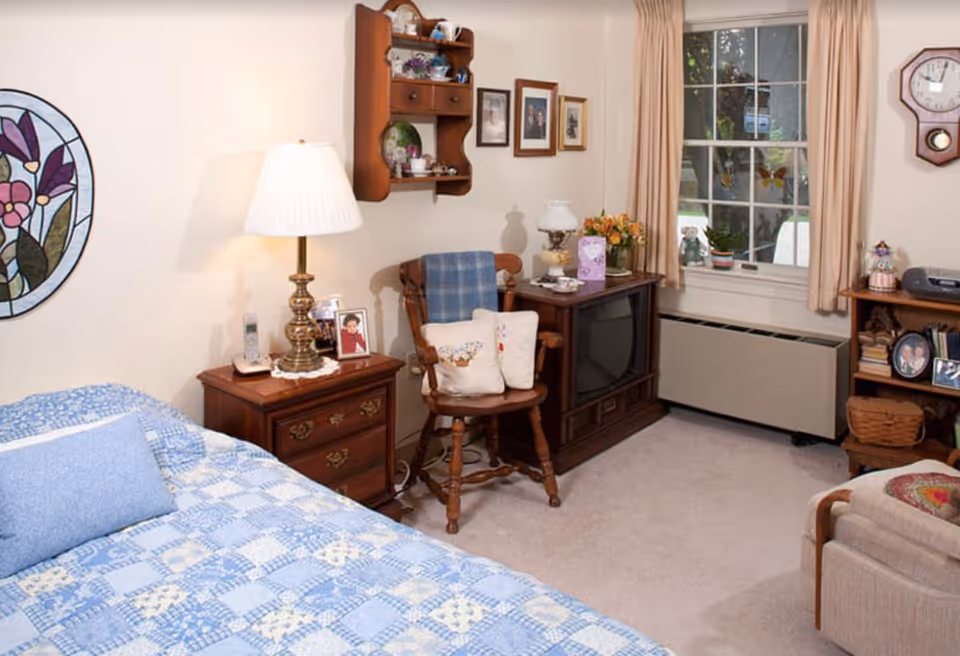 Cozy bedroom with a blue and white patchwork quilt on the bed, a wooden nightstand with a lamp and framed photos, a wooden chair with cushions, a wooden TV stand with an old-style television, a window with beige curtains, and various decorative items including a wall clock and framed pictures.
