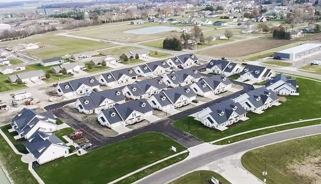 Aerial view of a residential complex of identical single-story villas with dark roofs arranged in neat rows amid roads and green lawns.