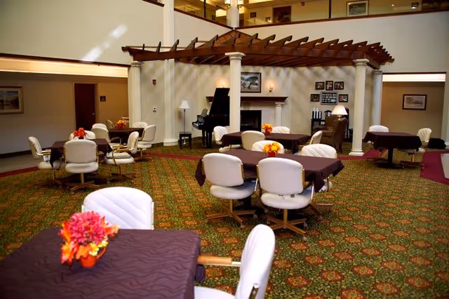 Spacious senior living dining area with round tables and white chairs, a fireplace and piano beneath a decorative wooden pergola.