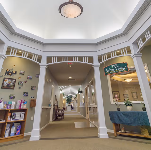 Bright interior corridor of a senior living facility with a central skylight, decorative archway, seating and an 'Arbor Village of Geneva Crossing' sign.