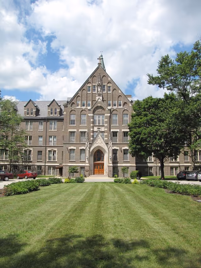Front exterior view of a large, multi-story brick building with a steeply pitched roof and arched entrance. The building is surrounded by a well-maintained lawn, trees, and shrubs under a partly cloudy sky.