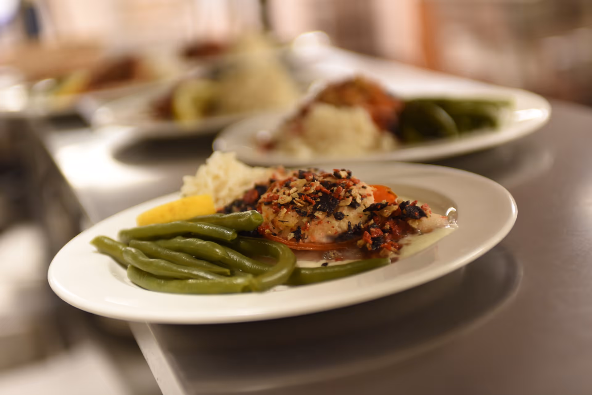 Plates of served meals on a stainless steel counter featuring green beans, rice and a topped fish portion.