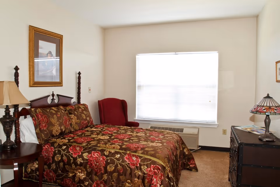 A furnished bedroom with a floral bedspread, bedside tables and lamps, a red armchair, and a window with blinds.