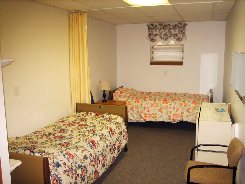 A small room with two single beds, each covered with floral bedspreads. There is a nightstand with a lamp between the beds, a small window with floral curtains, a chair, and a white dresser with a marble-like top. The walls are plain and the floor is carpeted.