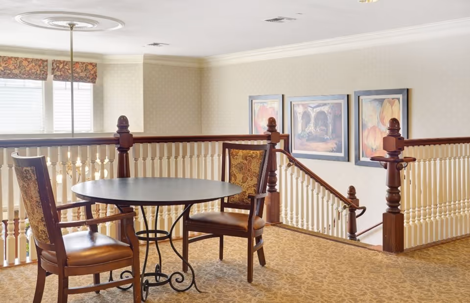 A small seating area with a round black table and two wooden chairs with patterned upholstery, situated near a staircase with white spindles and dark wooden handrails. The walls are light-colored with three framed paintings, and there is a window with floral valances letting in natural light.