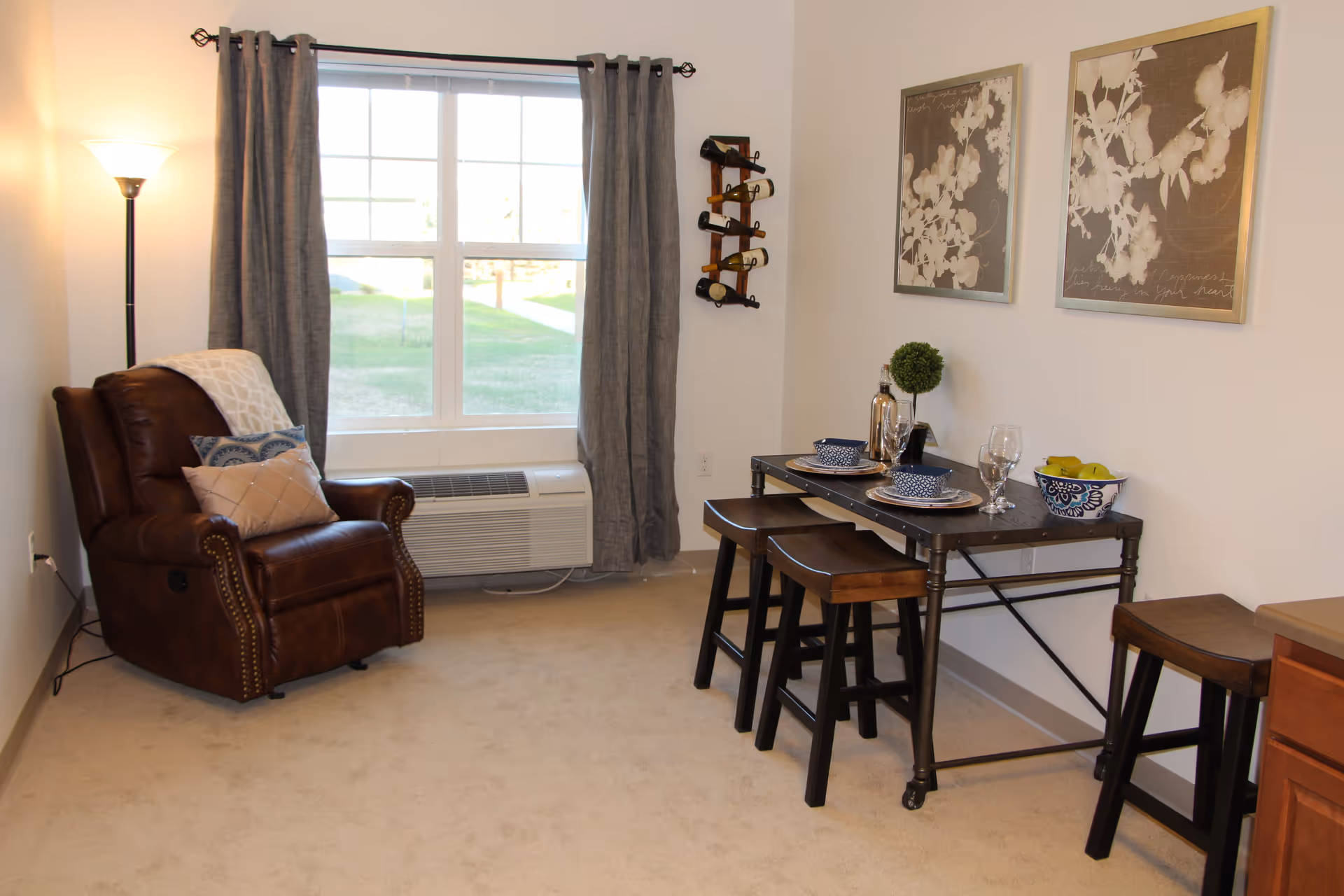 A cozy interior room with a brown leather recliner chair adorned with pillows and a throw blanket near a window with gray curtains. Next to the chair is a tall floor lamp. On the right side, there is a small dining table set with two place settings, a decorative plant, a bowl of fruit, and a wine bottle holder mounted on the wall with several bottles. Two framed floral artworks hang above the table.