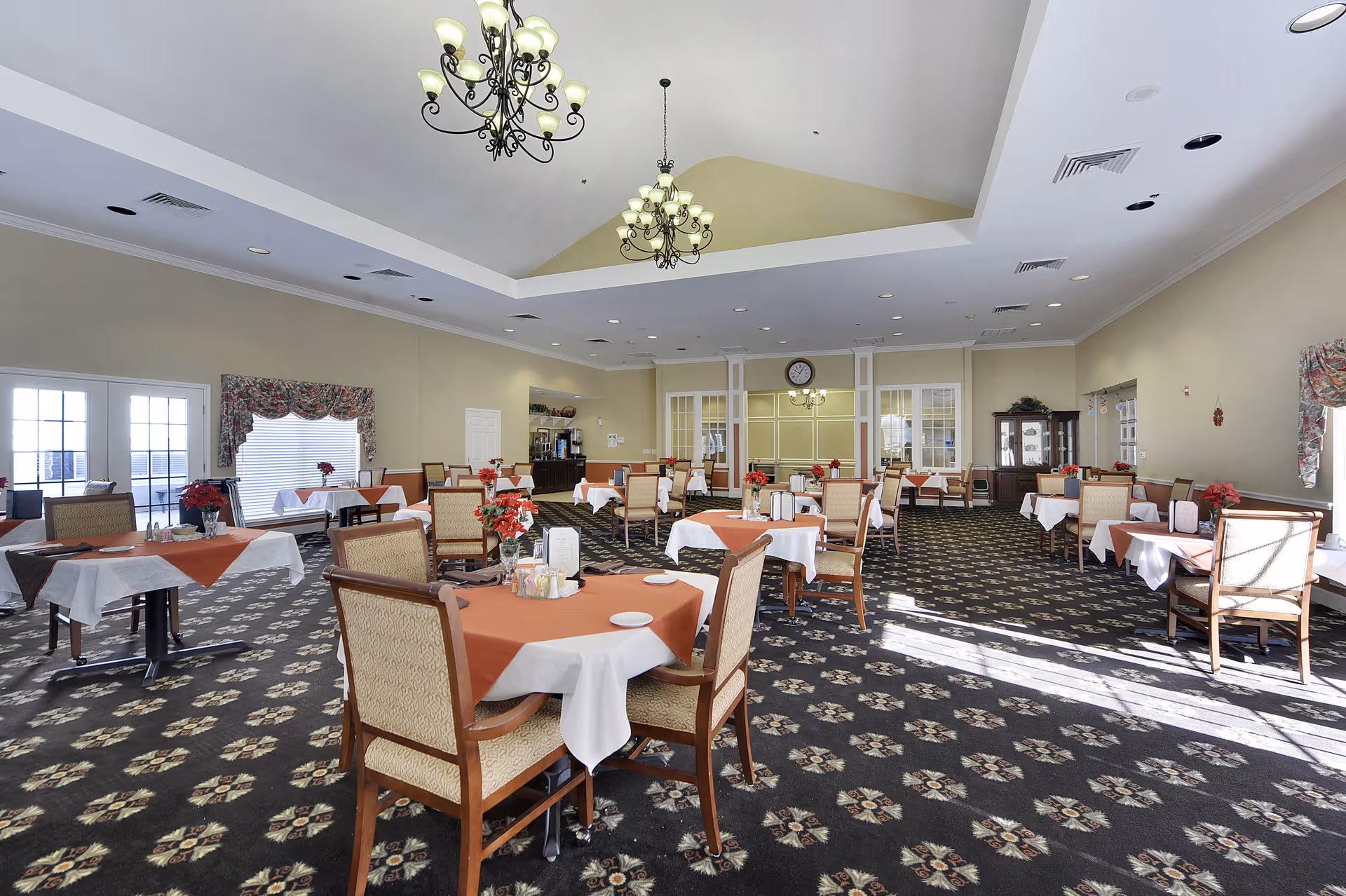 A spacious dining room with multiple tables covered in white and orange tablecloths, each set with chairs and small floral centerpieces. The room features patterned carpet, large windows with floral curtains, and two ornate chandeliers hanging from a high ceiling. There is a clock on the far wall above a set of glass-paneled doors.