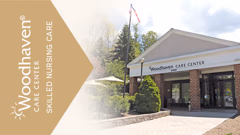 Entrance of Woodhaven Care Center building with a covered driveway, brick pillars, and a sign displaying the facility's name. There are trees and shrubs around the entrance and a flagpole with an American flag.