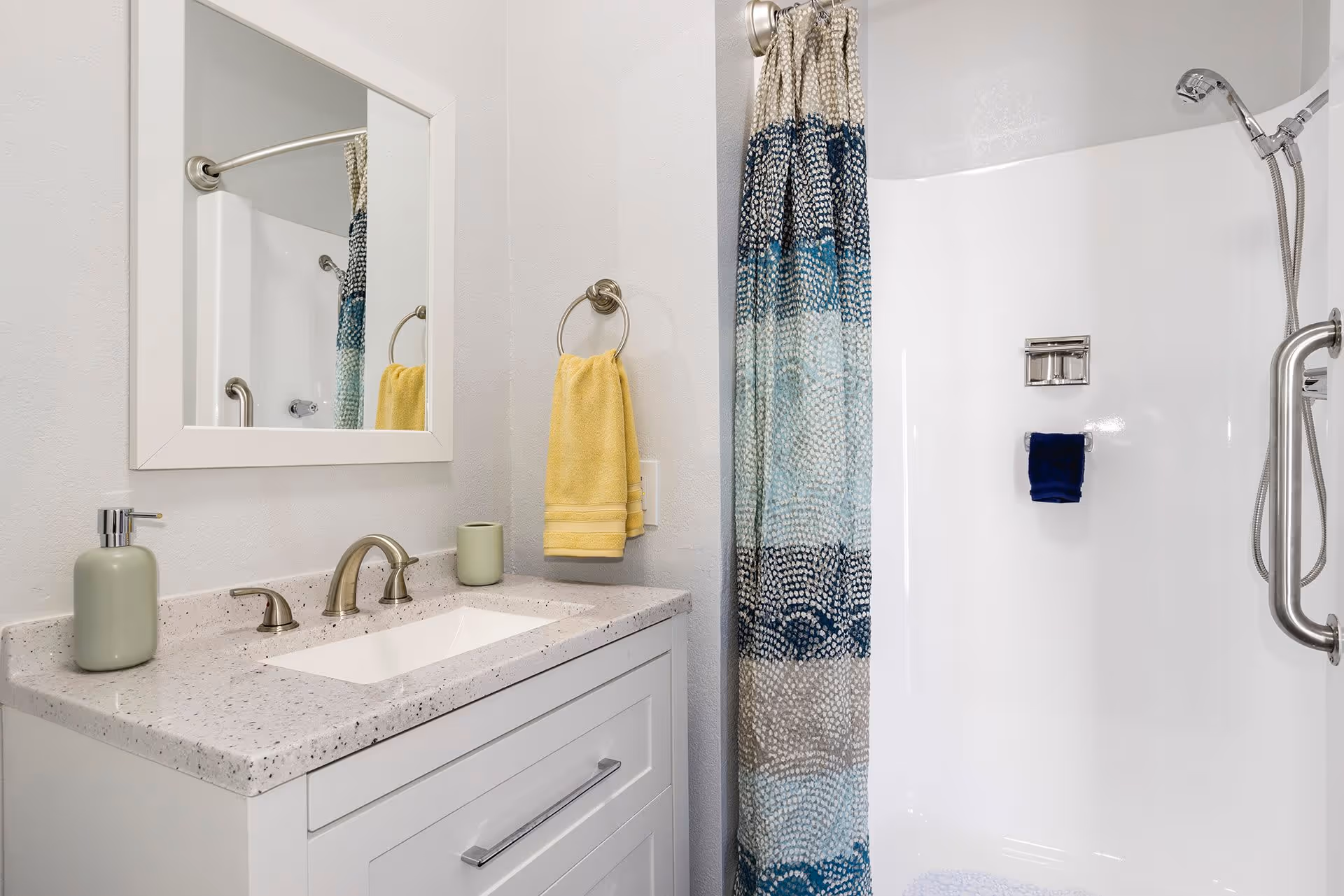 Small bathroom with a white vanity and speckled countertop, mirror and faucet on the left and a colorful shower curtain and walk-in shower with grab bar on the right.