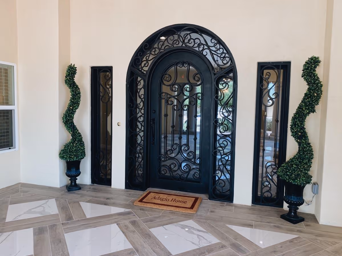 Decorative black wrought iron arched door with glass panels on either side, flanked by two spiral-shaped topiary plants in black pots, with a beige wall and patterned tile floor. A doormat in front of the door reads 'Adagio House'.